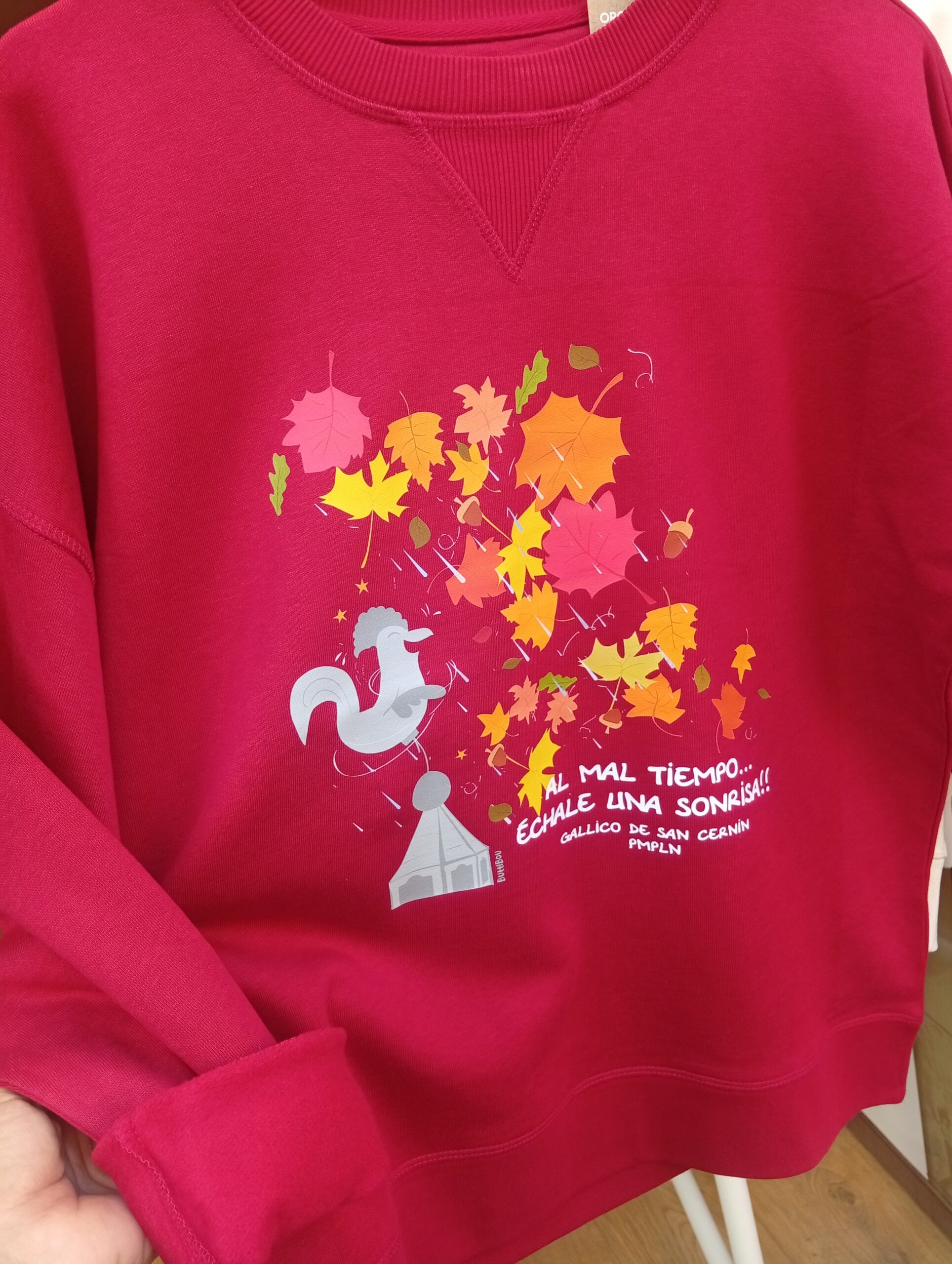 El espíritu del Gallico de San Cernin en una sudadera, con lluvia y hojas otoñales. Diseño ButtiBou Pamplona. Diseño propio de ButtiBou SudaderaGallico de San Cernin en color hibiscus red, diseño de ButtiBou Pamplona con hojas otoñales , lluvia y la frase " Al mal tiempo...échale una sonrisa"