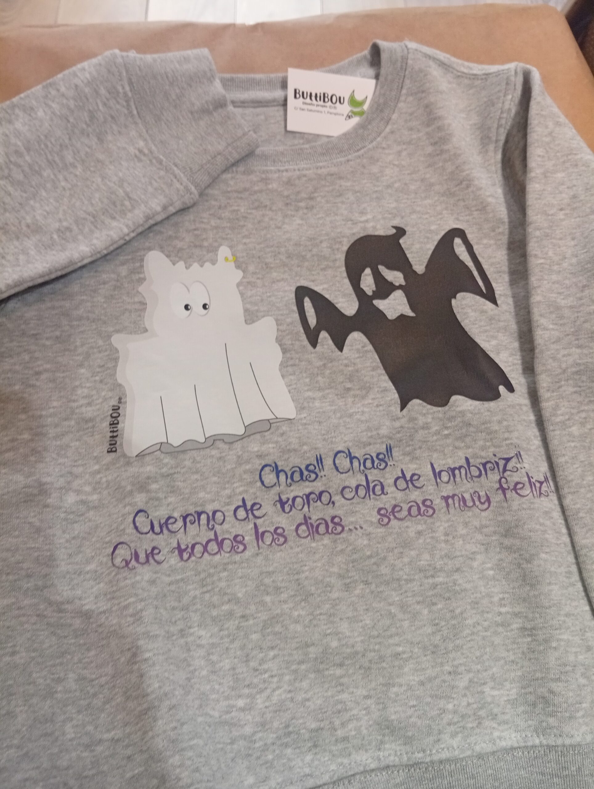 El pequeño becerrito de ButtiBou se disfraza de fantasma y te desea un Halloween lleno de alegría. The little ButtiBou calf dresses up as a ghost to wish you a happy and spooky Halloween!. Grey organic kids’ sweatshirt with ghost calf print and fun Halloween phrase “Chas chas! Bull’s horn, worm’s tail, may every day be happy” – exclusive design by ButtiBou Pamplona. Sudadera infantil gris orgánica con estampación de becerrito fantasma y frase divertida de Halloween “¡Chas chas! Cuerno de toro, cola de lombriz, que todos los días seas muy feliz”, diseño exclusivo de ButtiBou Pamplona.
