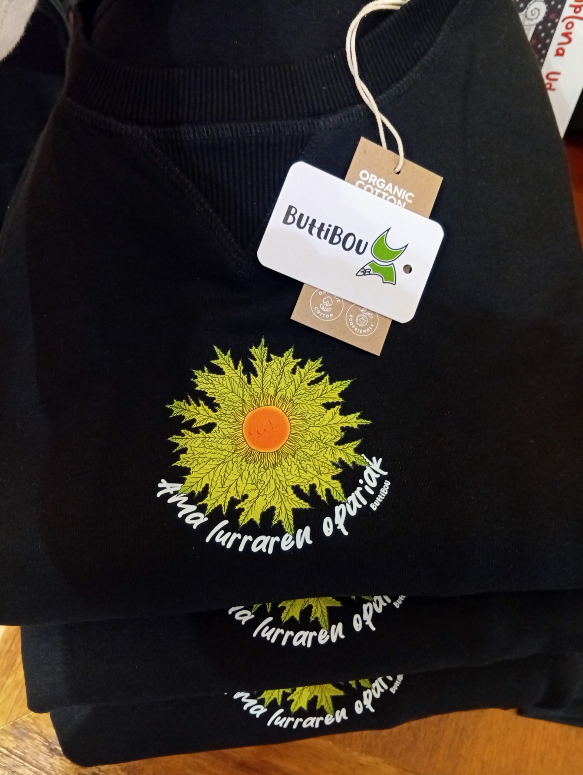 Sudadera oversize negra de mujer con eguzkilore verde y frase “Ama lurraren opariak”. Diseño exclusivo de ButtiBou Pamplona, comodidad y estilo inspirado en la tradición navarra. Black oversized women's sweatshirt with green eguzkilore and phrase “Ama lurraren opariak”. Exclusive design by ButtiBou Pamplona, comfort and style inspired by Navarre tradition. Sudadera oversize negra de mujer con eguzkilore verde lima y frase Ama lurraren opariak de ButtiBou Pamplona. Black oversized women's sweatshirt with green eguzkilore and phrase Ama lurraren opariak by ButtiBou Pamplona