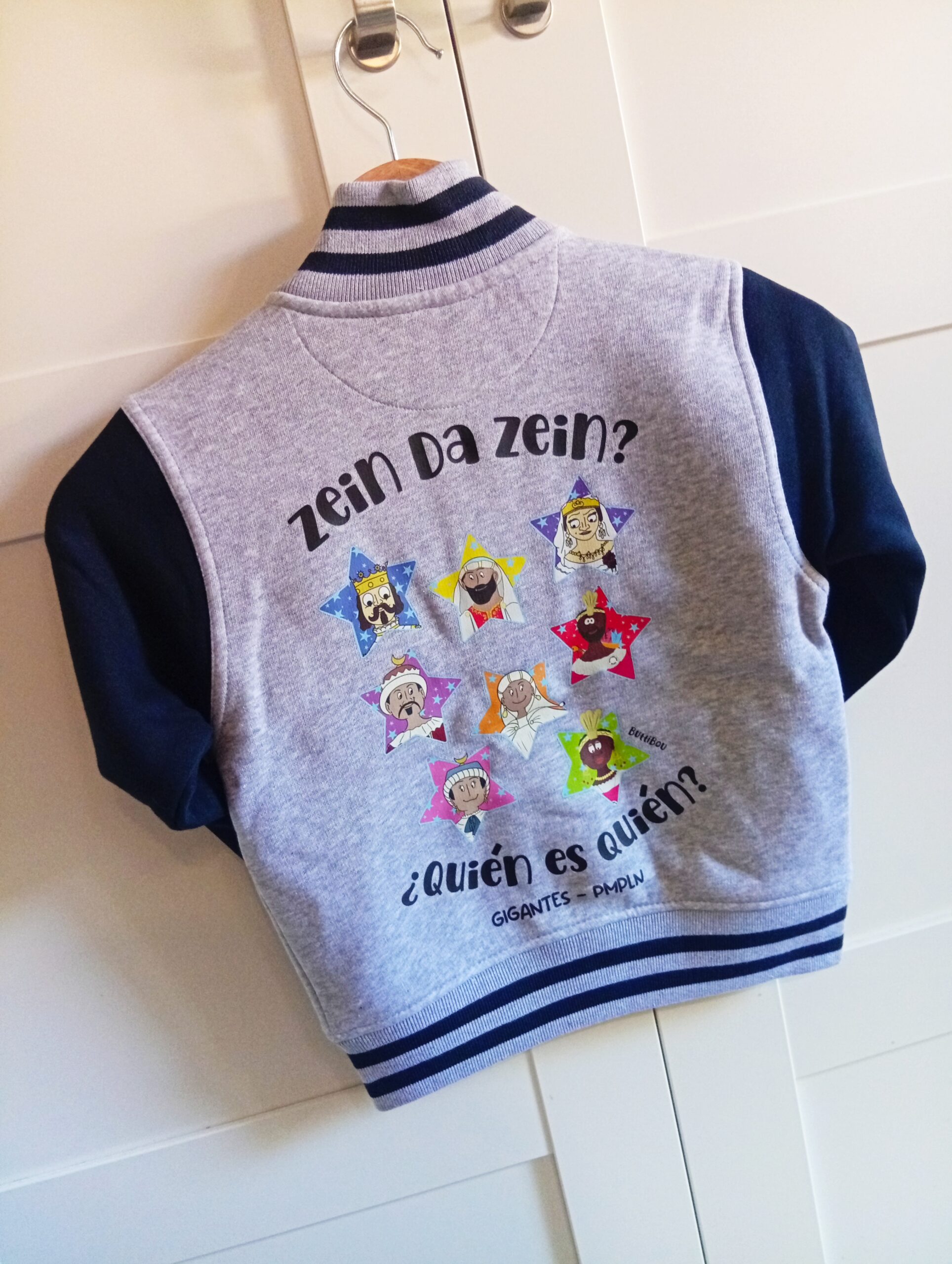 Sudadera estilo High School con interior gris claro, mangas azules en contraste y dobladillo, cuello y puños de canalé bicolor. Estampación espalda “Quién es Quién – Gigantes de Pamplona” y delantero “Gigantes JoseMiguelErico y JosePhamunda”. High School style sweatshirt with light gray interior, blue contrasting sleeves, and bicolor ribbed hem, collar, and cuffs. Back print “Quién es Quién – Pamplona Giants” and front print “Gigantes JoseMiguelerico and Josephamunda”. Sudadera High School con interior gris claro, mangas azules, dobladillo, cuello y puños de canalé bicolor. Estampación espalda “Quién es Quién – Gigantes de Pamplona” y delantero “Gigantes JoseMiguelErico y JosePhamunda”. High School sweatshirt with light gray interior, blue sleeves, bicolor ribbed hem, collar, and cuffs. Back print “Quién es Quién – Pamplona Giants” and front print “Gigantes JoseMiguelErico and JosePhamunda”.
