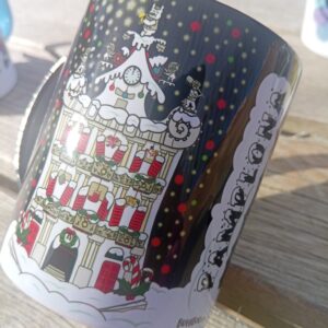 Taza de porcelana con interior negro y diseño navideño de la Plaza Consistorial de Pamplona decorada con nieve y luces, diseño exclusivo de ButtiBou Pamplona. Porcelain mug with black interior featuring a Christmas design of Pamplona’s City Hall Square decorated with snow and lights, exclusive ButtiBou Pamplona design.