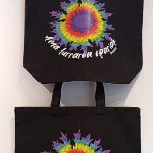 Totebag negro de algodón resistente con estampación Eguzkilore Pride cerca del centro y frase “Ama Lurraren Opariak”, diseño inclusivo y exclusivo ButtiBou Pamplona. Black cotton tote bag with Eguzkilore Pride print and the phrase “Ama Lurraren Opariak,” inclusive and exclusive design by ButtiBou Pamplona.