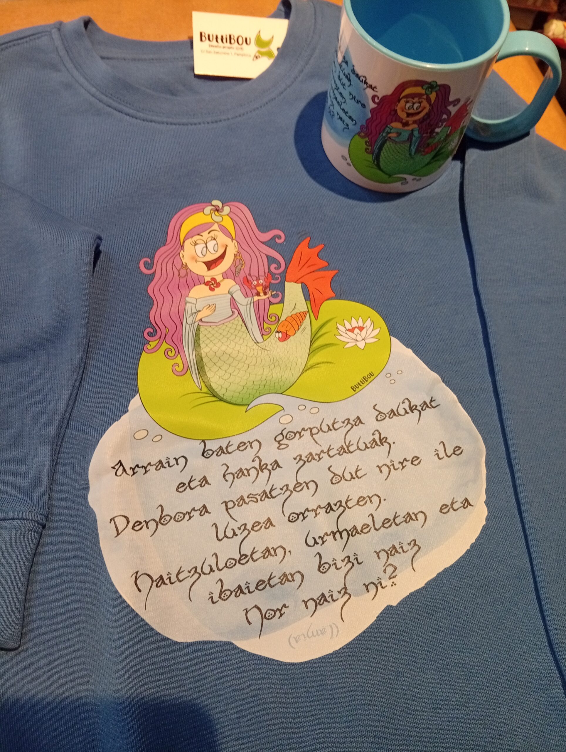 Una prenda mágica que conecta a los más pequeños con la mitologia vasca. A magical garment that connects little ones with the Basc mithologique Sudadera infantil orgánica azul con diseño de una lamia, figura mitológica vasca, y una adivinanza en euskera. Blue organic kids sweatshirt with a design of a lamia, Basque mythological figure, and a riddle in Basque.